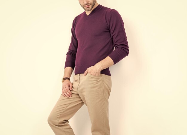A man in a deep purple sweater and beige trousers stands casually, hands in pockets, against a neutral background.