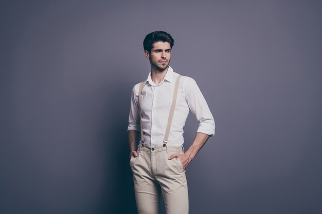 Man in white shirt and beige suspenders standing with hands in pockets against a plain gray background, looking confidently to the side.