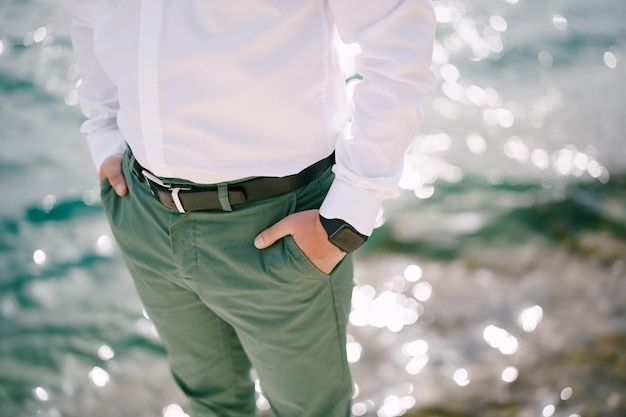 A man in a crisp white shirt and green trousers stands by the ocean, hands casually in his pockets.