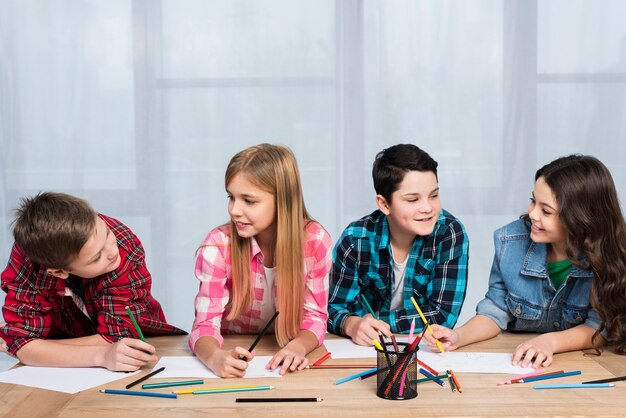 Group of children drawing with colored pencils at a table, smiling and collaborating during a classroom activity.